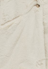 Tissu doux et pelucheux de couleur crème avec une couture de poche visible et un seul bouton beige près du bord supérieur.