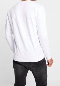 Hombre vestido con una camiseta blanca de manga larga y jeans oscuros, visto desde atrás sobre un fondo neutro.