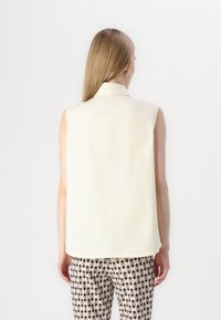 Camisa sin mangas amarilla con cuello, hecha de una tela ligera. La parte trasera tiene un corte recto y llega hasta las caderas. Combinada con pantalones de estampado.