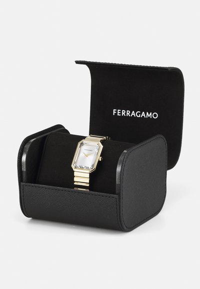 Kuldse tooni ristkülikukujuline kell valge näoga, millel on minimalistlik disain tekstuuritud musta ekraani ümbrises. Nähtav Ferragamo logo.