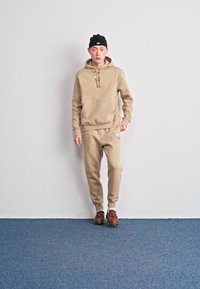 Hoodie bege e calças de treino coordenadas, feitas de um tecido suave, complementadas com sapatos castanhos. Design simples com um pequeno logotipo do lado esquerdo.