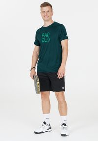 Mörkgrön träningsskjorta med texten "PAD" och "EL", svarta shorts och vita strumpor. Håller en padelracket och står framför en enfärgad bakgrund.