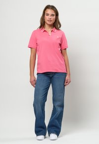 Mlada ženska stoji, oblečena v roza polo majico, modre široke kavbojke in bele superge na enostavni svetli podlagi.