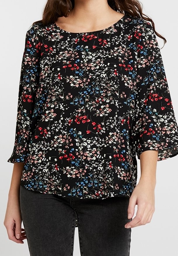 Kvinna klädd i en svart blommig blus med röda, rosa och blå blommor, i kombination med mörka jeans och avslappnade ärmar.