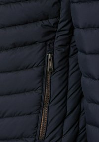 Nära bild av mörk vadderad jacka med en synlig svart dragkedja på sidofickan, som visar quiltade horisontella sömmar.