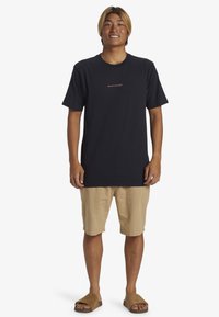 Camiseta de algodón negra con un pequeño logo naranja, pantalones cortos beige y sandalias marrones. Ajuste relajado con manga corta y cuello redondo.