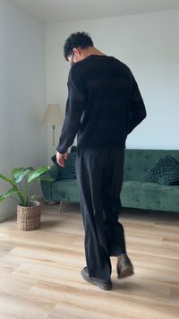 Fekete, bő szabású pulóver texturált csíkokkal, párosítva széles szárú fekete nadrággal és sötét cipőkkel, egy világos szobában, ahol növény és kanapé is található.