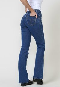 Jeans de mezclilla azules ajustados con pernera acampanada, con bolsillos traseros cosidos y una cintura de altura media, contra un fondo neutro.
