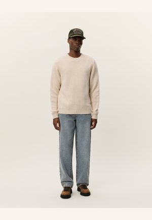 Beige knit sweater, light blue jeans, and brown shoes. A green cap with white lettering completes the outfit, emphasizing casual attire.