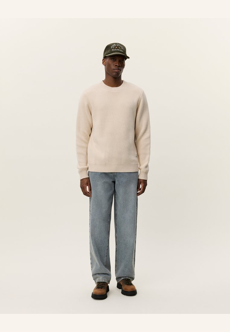 Beiger Strickpullover, hellblaue Jeans und braune Schuhe. Eine grüne Mütze mit weißer Schrift vervollständigt das Outfit und betont die legere Kleidung.