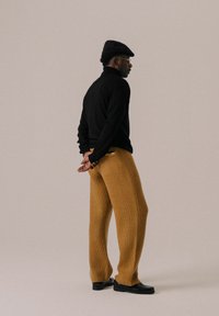 Maglione nero a maglia, pantaloni texturizzati color senape, berretto piatto nero e scarpe nere, visti di lato su uno sfondo neutro.