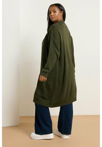 Lång olivgrön stickad cardigan med en lös passform, strukturerat mönster och ribbad nederkant. Kombinerad med mörkblå vida jeans och vita sneakers.