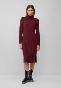 Bordeaux-farbenes Langarmkleid mit hohem Hals und seitlichem Schlitz, gefertigt aus einem weichen Material, kombiniert mit schwarzen derb-sohlen Knöchelstiefeln.