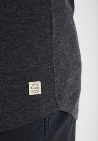 Tmavě šedé tričko s jemnou texturou, na dolní straně s obdélníkovou krémovou nášivkou s nápisem "DENIM BLEND WITH ATTITUDE".
