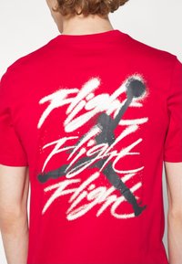 Punainen puuvillapaita, jossa on suuri musta siluetti hyppivästä hahmosta, ja selässä valkoisella graffiti-tyylillä kirjoitettu sana "Flight".