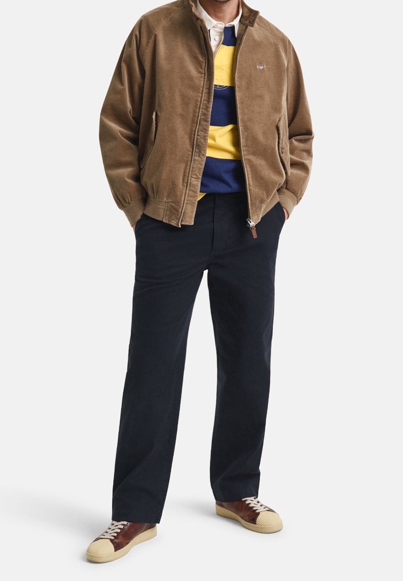Homme portant une veste marron à fermeture éclair sur une chemise rayée jaune et marine, un pantalon foncé et des baskets marron et beige, debout les mains dans les poches.