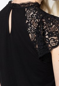 Svart topp med spetsärmar med blommönster. Nyckelhålsdetalj på baksidan ger en unik designfunktion. Slät tygtextur.