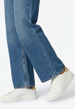 Legs wearing blue straight-leg jeans and white low-top sneakers with beige heel detail, against light background.