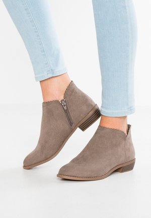 Personne portant un jean skinny bleu clair et des bottines en daim taupe avec fermetures éclair latérales et bords festonnés.