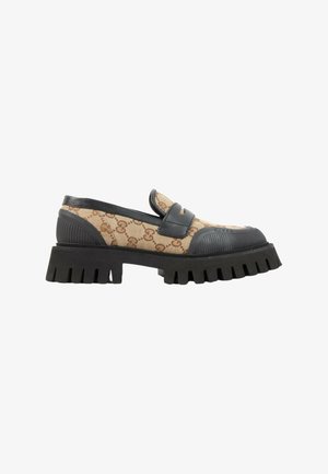 Chaussure mocassin avec tissu beige à motifs, accents en cuir noir et semelle épaisse en caoutchouc noir à relief, vue de côté sur fond blanc.