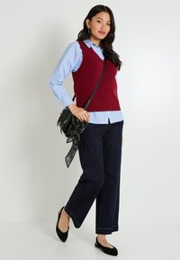 Femme portant un pull sans manches rouge sur une chemise bleue, un jean large noir, des ballerines noires et portant un sac bandoulière noir avec une écharpe.