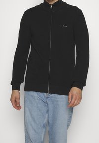 Man som bär en svart strukturerad zip-up-tröja med en vit GANT-logotyp på bröstet och ljusblå jeans, stående mot en enfärgad bakgrund.
