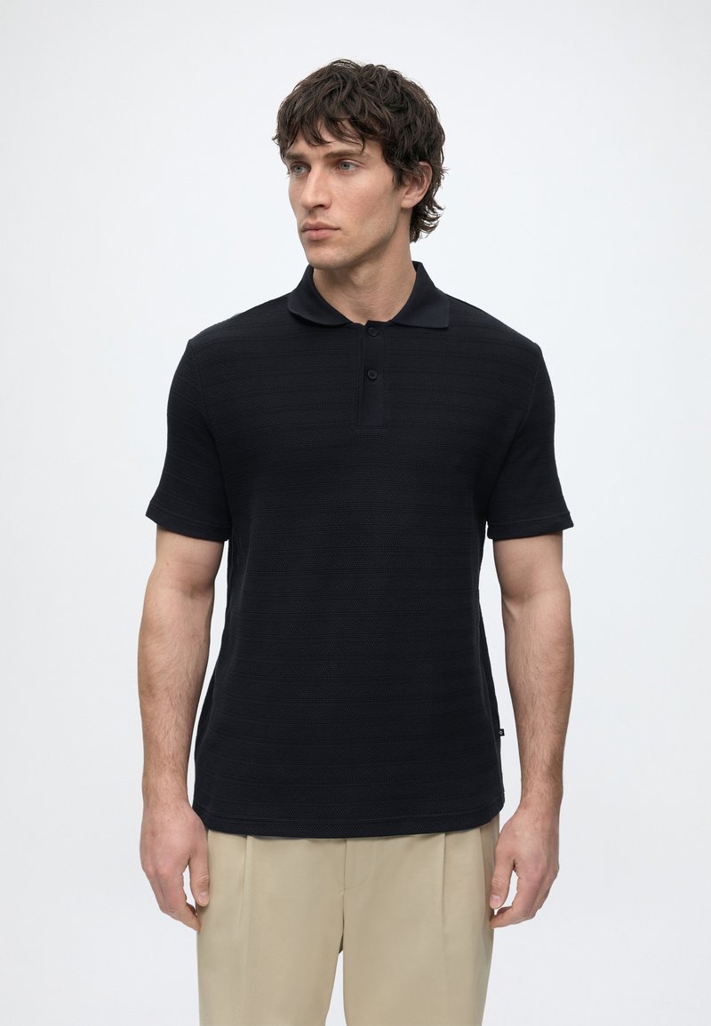 Homme debout sur un fond uni, portant un polo noir à manches courtes et un pantalon beige, regardant légèrement vers la gauche.