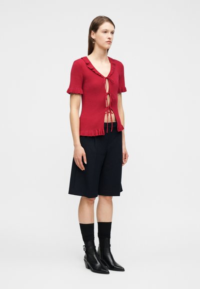 Mujer joven con un top de punto rojo de manga corta con lazadas frontales, pantalones cortos negros hasta la rodilla, calcetines negros y botines negros, de pie de perfil.