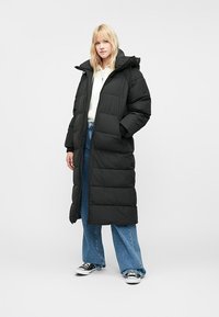 Nő hosszú, fekete steppelt kabátban, kapucnival, fehér pulcsiban, bő kék farmernadrágban, és fekete-fehér sportcipőben áll, kezével a zsebében.