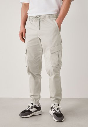 Lysegrå cargobukser med elastisk talje og manchetter, sidelommer, båret med sorte og hvide sneakers og en hvid t-shirt.