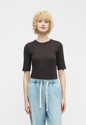 Femme aux cheveux blonds courts et bouclés portant un haut noir ajusté avec des manches aux coudes et un pantalon en denim bleu clair ample avec des cordons blancs.