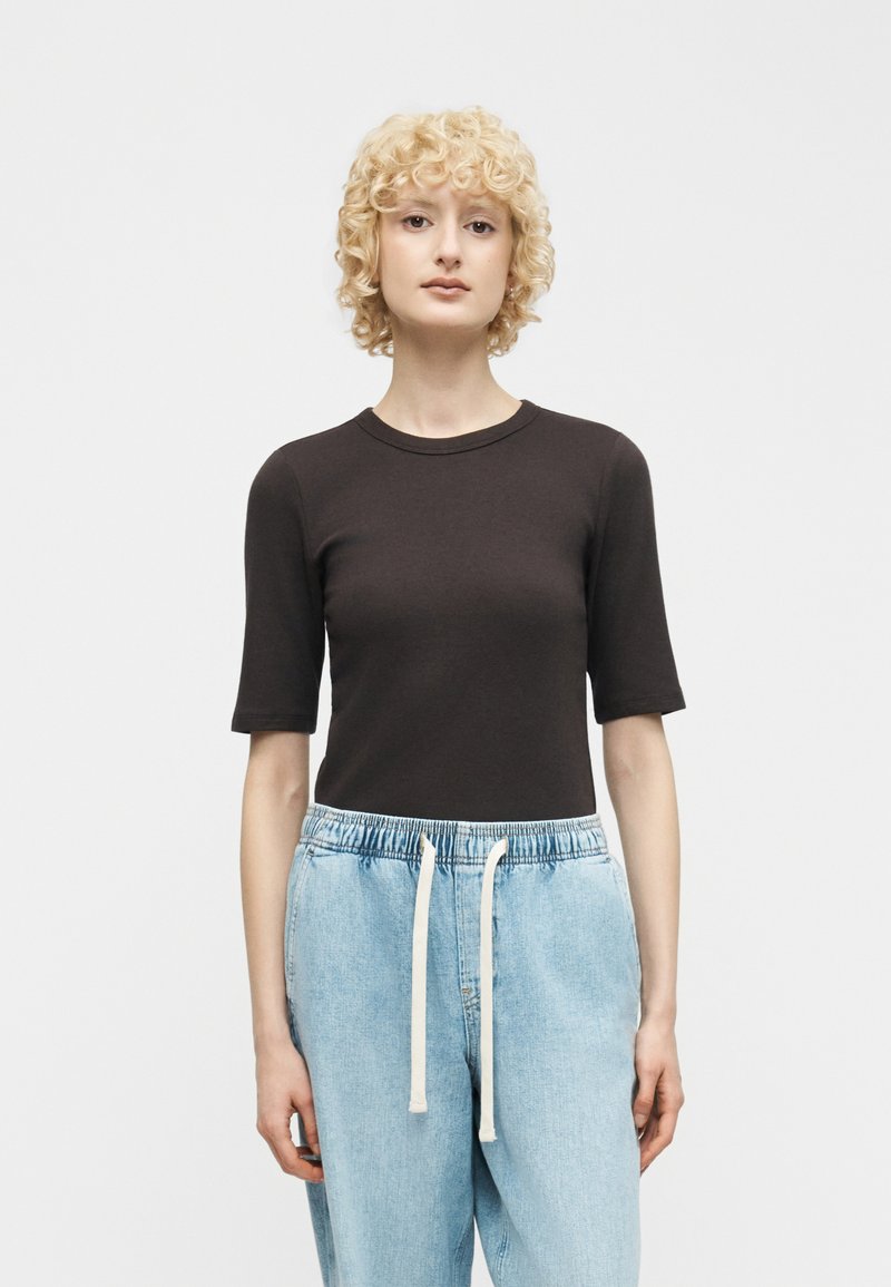 Γυναίκα με κοντά σγουρά ξανθά μαλλιά, φορώντας μαύρο εφαρμοστό τοπ με μανίκια έως τον αγκώνα και ανοιχτόχρωμο μπλε τζιν παντελόνι με άσπρα κορδόνια.