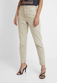 Jeans de pierna recta de talle alto en color beige con una textura ligera, cinco bolsillos y pasadores para cinturón, combinados con unos tacones altos negros con tiras.