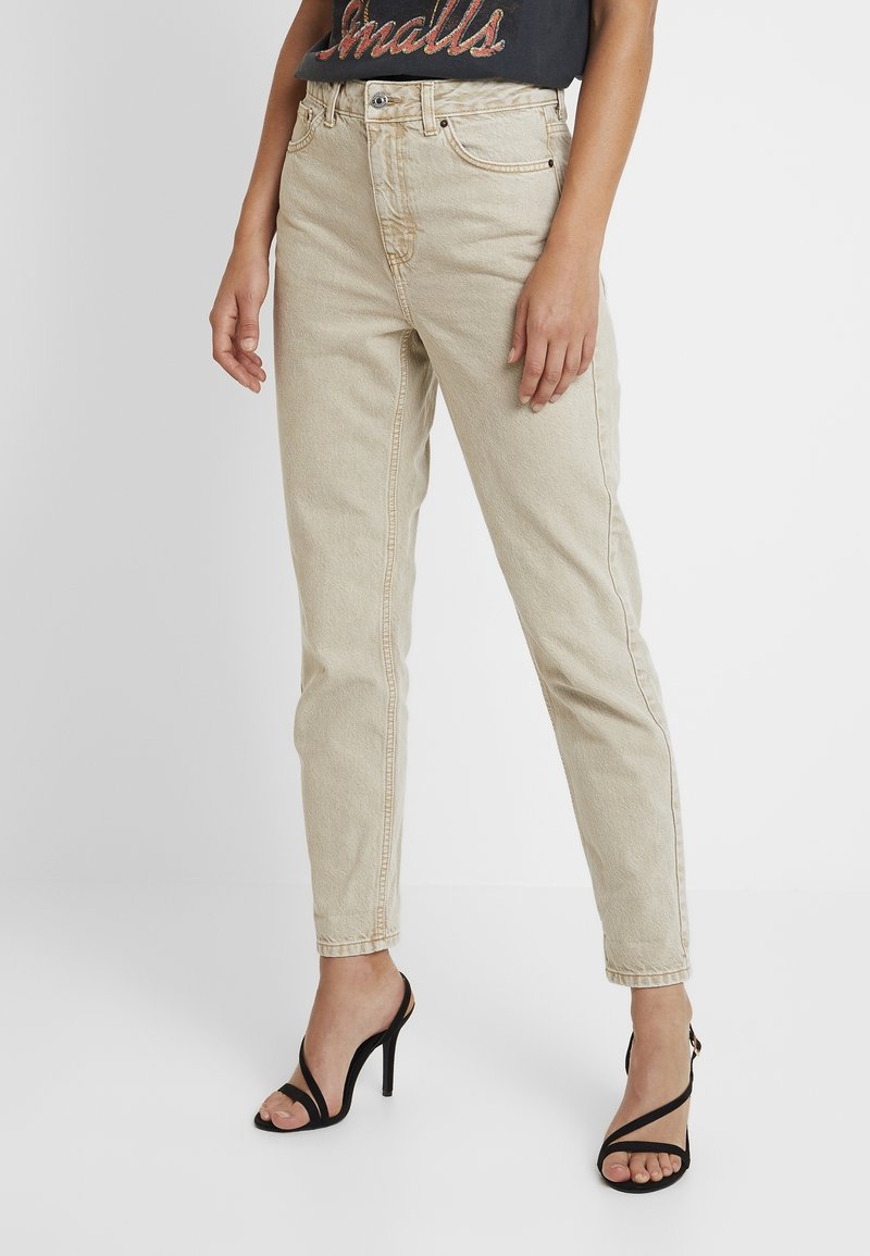 Jeans de pierna recta de talle alto en color beige con una textura ligera, cinco bolsillos y pasadores para cinturón, combinados con unos tacones altos negros con tiras.