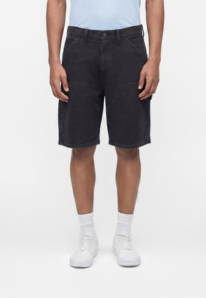 Uomo che indossa pantaloncini neri fino al ginocchio con tasche frontali, calzini bianchi e scarpe da ginnastica bianche, in piedi davanti a uno sfondo bianco semplice.