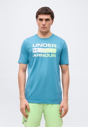 Mladý muž v modrom tričku značky Under Armour a svetlozelených šortkách stojaci pred jednoduchým bielym pozadím.