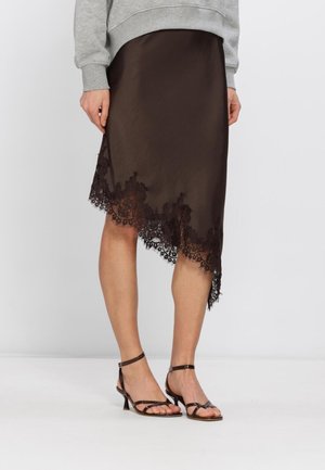 Femme portant une jupe asymétrique marron avec des bordures en dentelle, des talons noirs à brides et un haut gris à manches longues, debout sur un sol blanc.