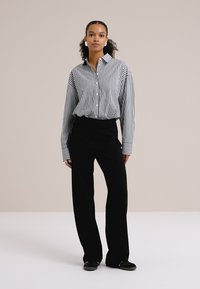 Oseba samozavestno stoji v prugasti srajci na gumb in širokih črnih hlačah pred nevtralno ozadje, s čimer prikazuje moden in stilski videz.