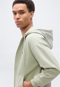 Νεαρός άνδρας με ανοιχτό πράσινο φούτερ με φερμουάρ και τα χέρια στις τσέπες, κοιτάζοντας λίγο πάνω από τον ώμο του μπροστά σε έναν απλό φωτεινό φόντο.