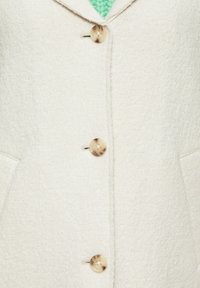 Manteau couleur crème en tissu texturé, avec un col à revers, cinq boutons et des poches latérales. Les boutons ont une finition marbrée.