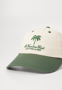 Baseballkasket med en cremefarvet krone og grøn skygge. Har en grøn broderet palmetræ og teksten "On Vacation Club LET'S TAKE A TRIP."
