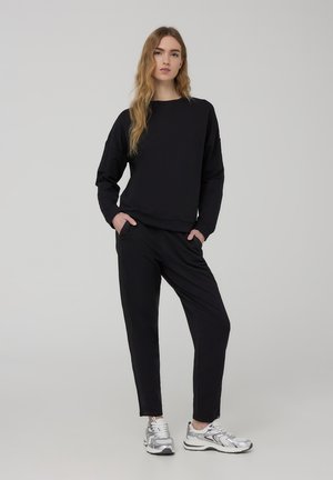 Czarny bawełniany sweatshirt z długimi rękawami, o luźnym kroju, zestawiony z czarnymi spodniami wąskimi. Model nosi srebrne buty sportowe. Proste tło.