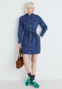 Denim shirt dress with long sleeves, collar, button front, two chest pockets, and a tie waist. Worn with green sneakers and a brown bag.