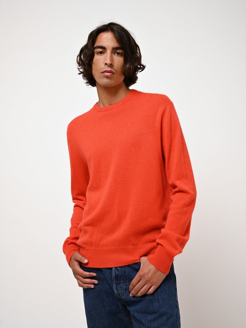 En orange ulltröja med rund halsringning och ribbade muddar, matchad med blå denimjeans. Tyget verkar mjukt med en slät textur.