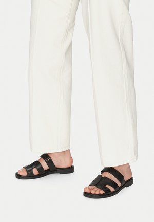 Black leather sandals with open toe design, featuring multiple straps and a flat sole. Worn with white, loose-fitting trousers.