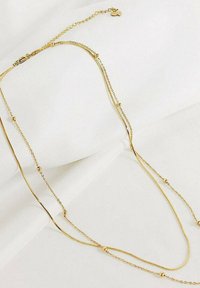 Collar de oro laminado con varios estilos de cadena, presenta cuentas redondas y un cierre de langosta sobre un fondo de tela blanca.