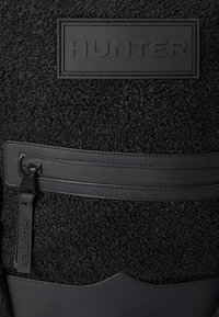 Černý látkový batoh s texturovaným, huňatým povrchem. Obsahuje černou gumovou kapsu na zip s viditelným logem "HUNTER" a hladkými akcenty.