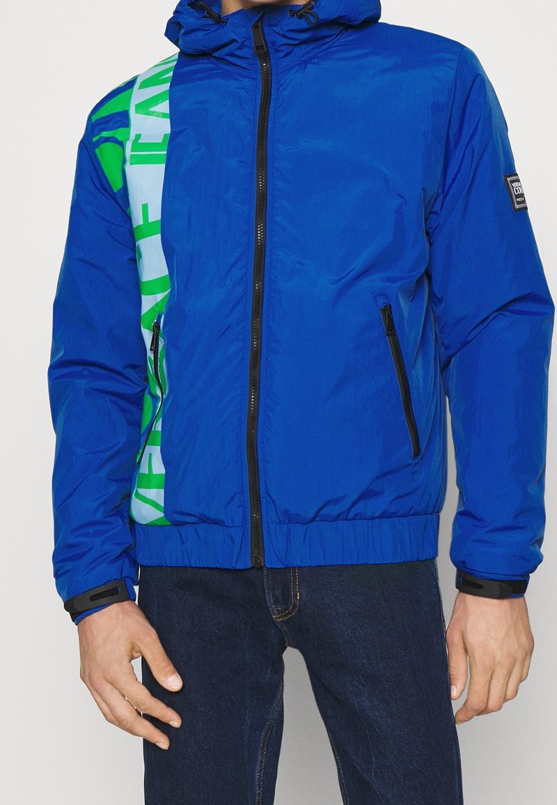 Blå vadderad jacka med huva, med gröna och vita grafiska ränder. Framsidan har dragkedja och två sidofickor med svarta dragkedjor.