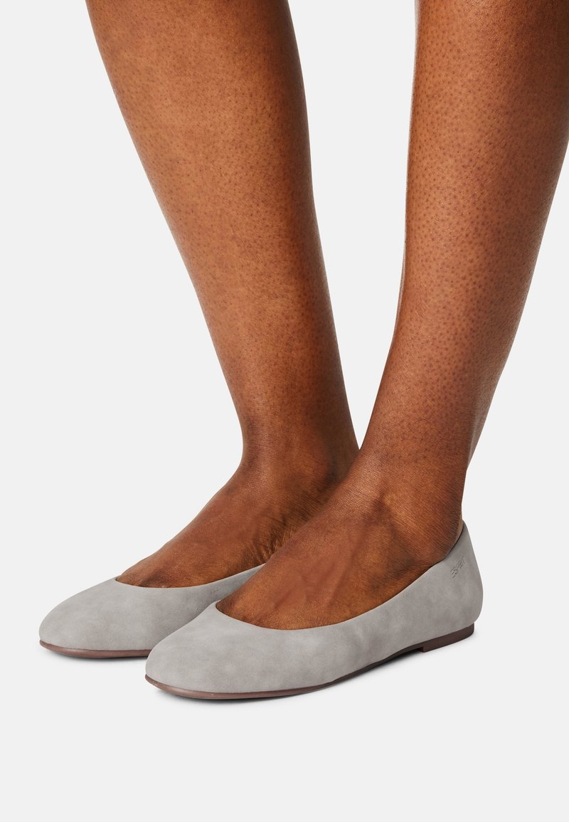 Pieds portant des ballerines en daim gris clair avec des bouts arrondis et un petit talon, sur un fond blanc uni.