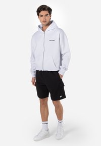 Hvid zip-up hoodie med frontlommer og sort logo; parret med sorte cargo shorts og hvide sneakers. Enkel design, afslappet pasform.